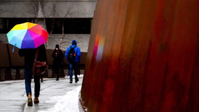 student walking by a wooden structure holding a rainbow umbrella