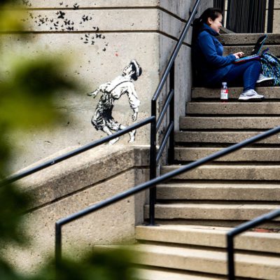 A student works on a laptop next to graffiti created by Jef Aerosol, an internationally renowned stencil artist, on the steps of the Richardson Plaza clock tower