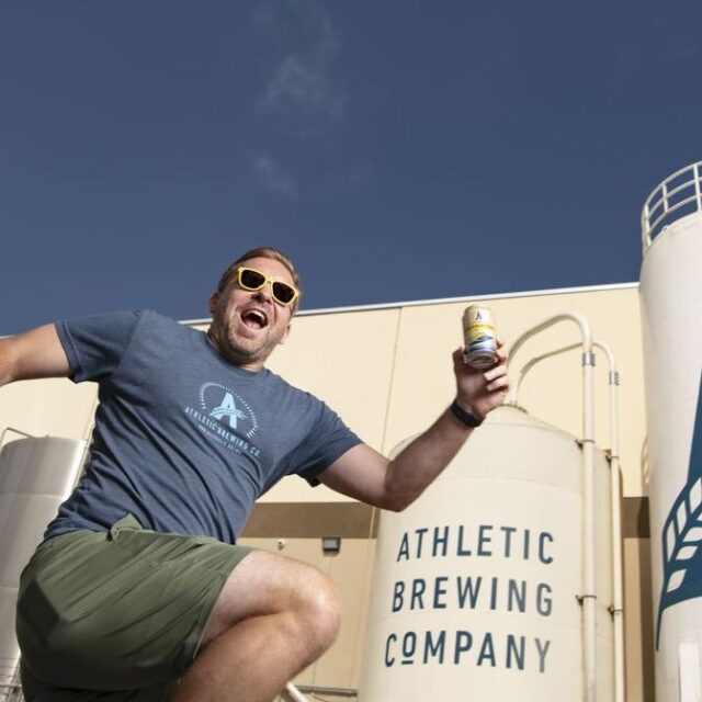 Athletic Brewing founder Bill Shufelt outside the Connecticut-based company’s new brewery in San Diego. Shufelt is wearing yellow sunglasses and holding a can of beer while exuberantly jumping in front of the brewery in the background.