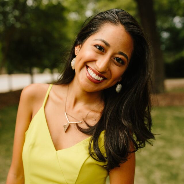 headshot of Genny. She is wearing a yellow dress, earrings, and a necklace and smiling.