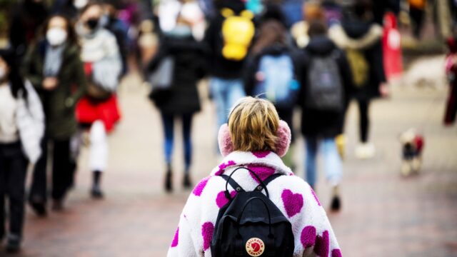 a student wearing a black backpack, pink earmuffs, and a pink fluffy jacket covered in hot pink hearts walks through a crowd of people outside on campus