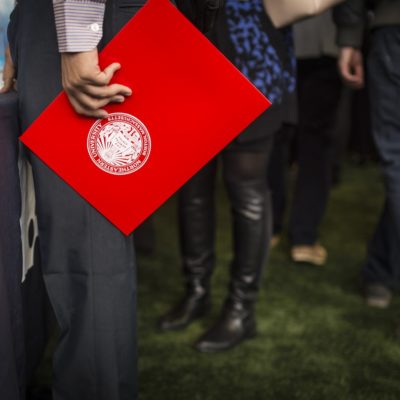 Zoomed in image of a person holding a red Northeastern folder standing on green grass in front of a black table.