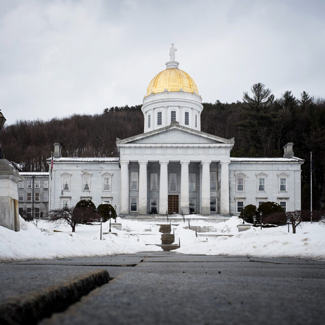 The gold domed Vermont State House in the capital city of Montpelier on a snowy day in winter time. There is snow all over the ground.