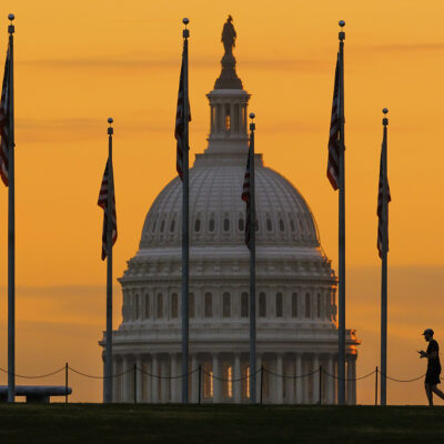 Photograph of the white house at sunset, with American flags lined in front of it. A person walks in front of them.