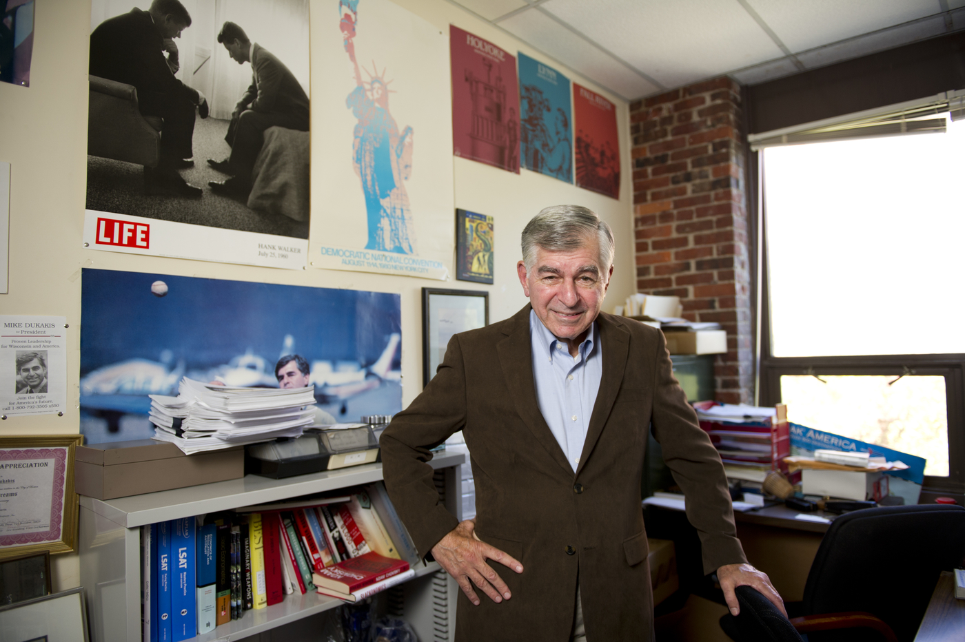 Picture of Professor Michael Dukakis in his office at Northeastern University. He is smiling and surrounded by posters and books on modern political science.