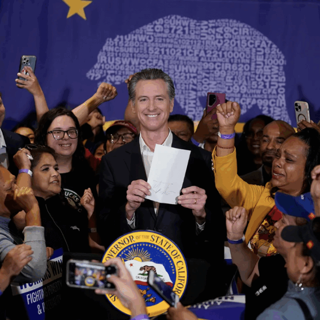 image of California Governor Gavin Newsom holding bill signing new minimum wage into law with people surrounding him emotional