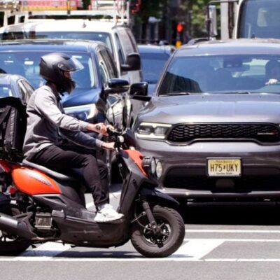 image of a delivery driver on a scooter rides the pedestrian crosswalk through traffic on a delivery in the Seaport District, Friday, June 7, 2024, in Boston.