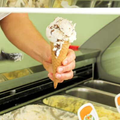 Female hand is holding a vanilla with dried fruit ice cream in waffle cone. Ice cream fridge with steel service containers in background. Worker in ice cream shop.