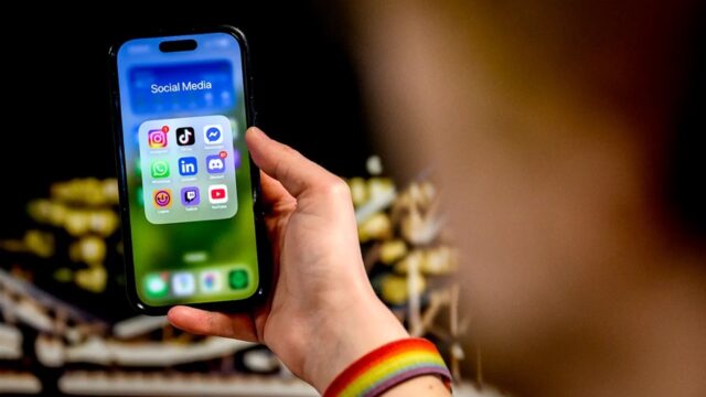 A hand holds up an iPhone with social media apps on the screen.