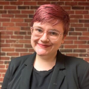 headshot of Juniper Johnson. They are wearing a black blazer and clear pink glasses. They have rose-colored short hair.