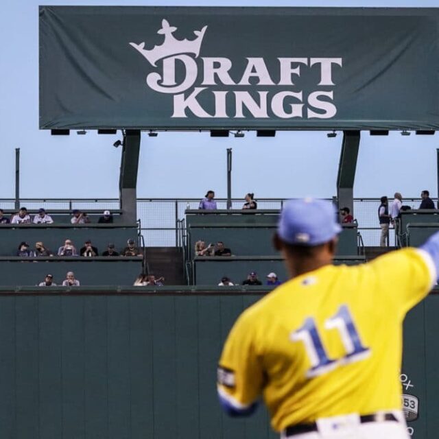 A left field scoreboard is covered under a DraftKings promotional as Boston Red Sox third baseman Rafael Devers (11) warms up before a baseball game at Fenway Park, Wednesday, Sept. 22, 2021, in Boston.