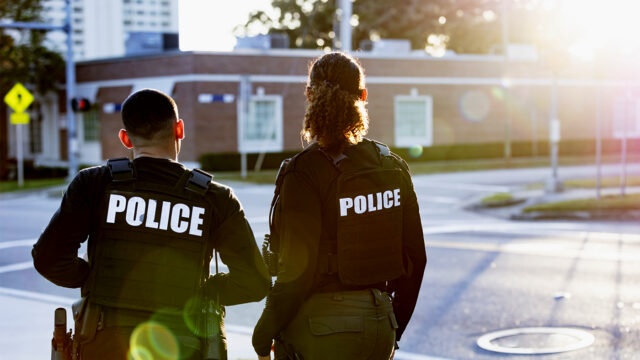 Rear view of two multiracial police officers patrolling a community on foot. They are standing at a street corner looking toward an empty intersection. The policewoman is mixed race, African-American, Asian and Hispanic, in her 40s. Her partner is a young Hispanic man in his 20s.