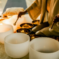 A woman in a calm space plays crystal and metal sound bowls surrounded by candles and lights, symbolizing feminine energy, relaxation, mindfulness, and healing meditation at a retreat.