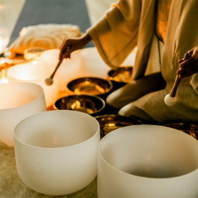 A woman in a calm space plays crystal and metal sound bowls surrounded by candles and lights, symbolizing feminine energy, relaxation, mindfulness, and healing meditation at a retreat.