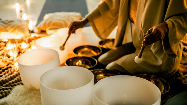 A woman in a calm space plays crystal and metal sound bowls surrounded by candles and lights, symbolizing feminine energy, relaxation, mindfulness, and healing meditation at a retreat.